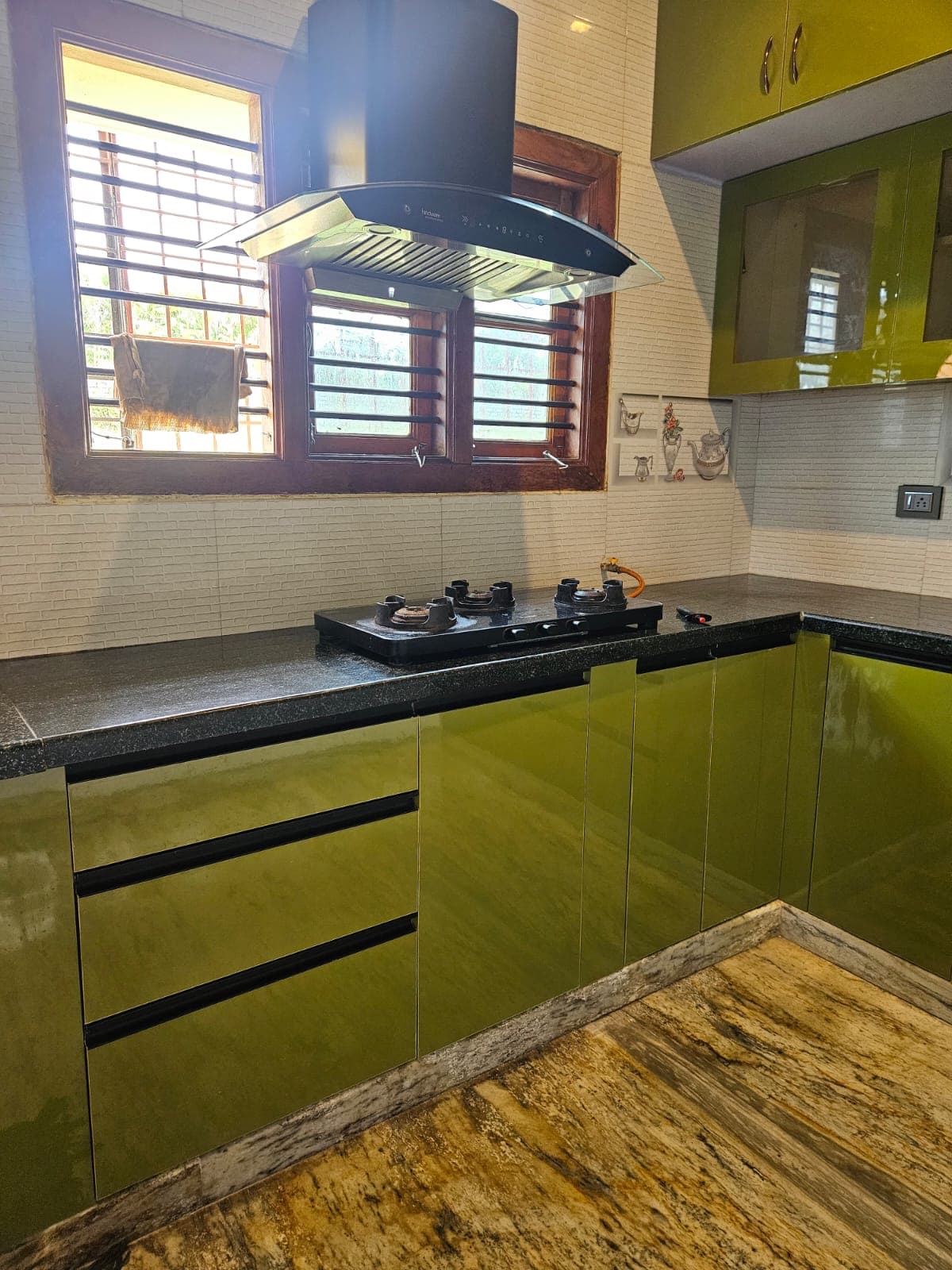 L-shaped modular kitchen in olive-green high-gloss shutters with a black granite counter and glass-fronted wall units